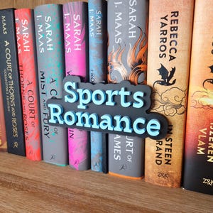 Op de afbeelding: Een boekenkast met een verscheidenheid aan boeken, waaronder "A Court of Thorns and Roses" en "Mist and Fury" van Sarah J. Maas, en "A Court of Wings and Ruin" van Sarah J. Maas. De boekenkast heeft ook een bord met de tekst "Sports Romance" in blauwe en zwarte letters.