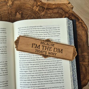 Könnte beinhalten: Rechteckiger Holz-Lesezeichen mit dem Text "BECAUSE I'M THE DM THAT'S WHY". Das Lesezeichen hat dekorative Ecken und liegt auf den Seiten eines offenen Buches. Der Text ist in einer Serifenschrift.