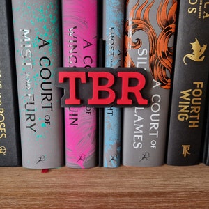 May include: A red and black bookend with the letters "TBR" in red. The bookend is sitting on a bookshelf with a row of books. The books have titles like "A Court of Thorns and Roses", "A Court of Mist and Fury", "A Court of Wings and Ruin", "A Court of Frost and Starlight", "A Court of Silver Flames", "Fourth Wing", and "Flame"