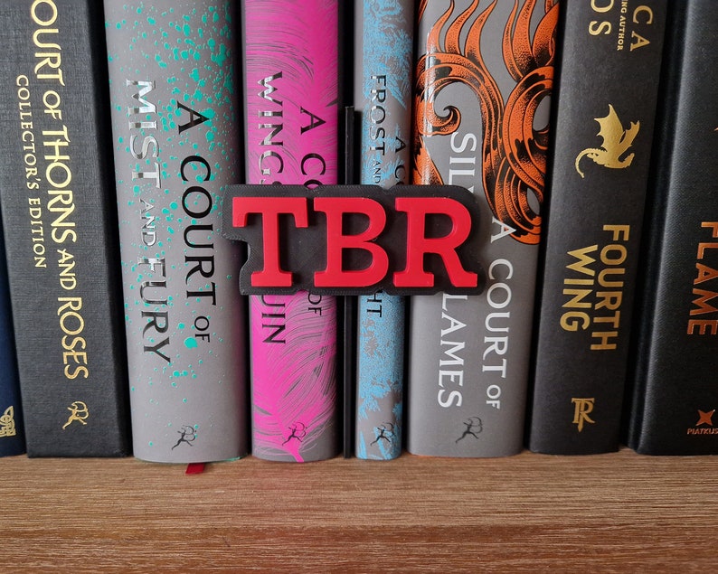 May include: A red and black bookend with the letters "TBR" in red. The bookend is sitting on a bookshelf with a row of books. The books have titles like "A Court of Thorns and Roses", "A Court of Mist and Fury", "A Court of Wings and Ruin", "A Court of Frost and Starlight", "A Court of Silver Flames", "Fourth Wing", and "Flame"