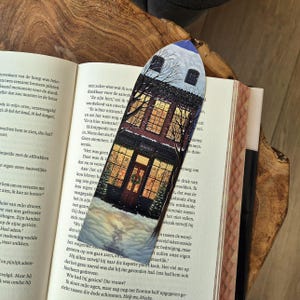 Op de afbeelding: Een decoratieve boekenlegger met een gedetailleerde illustratie van een boekwinkel 's nachts, met besneeuwde daken en een warme gloed van de ramen. Het woord "BOOKS" is zichtbaar boven de deur. De boekenlegger ligt in een open boek.