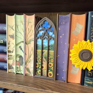 May include: A brown, arched, gothic-style book nook insert featuring a miniature landscape of a dirt path winding through a field of yellow sunflowers under a blue sky, positioned between several decorative books with floral and celestial designs.