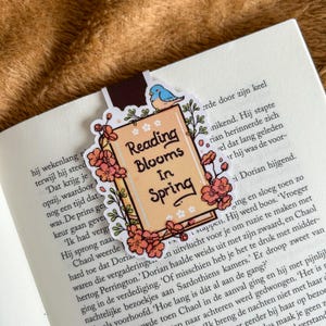 Op de afbeelding: Een decoratieve boekenlegger met een boekontwerp en de tekst "Reading Blooms In Spring". De boekenlegger is versierd met roze bloemen, groene bladeren en een blauwe vogel. Het ligt op de pagina's van een boek.