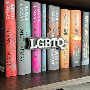 Op de afbeelding: Een houten boekenplank toont een verzameling fantasyromans met kleurrijke ruggen. Een wit en grijze "LGBTQ+" boekensteun staat voor de boeken. Titels zijn onder meer "A Court of Thorns and Roses" en "A Mist of Fury."