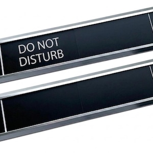 Könnte beinhalten: Zwei rechteckige Türschilder mit schwarzem Hintergrund und silberfarbenen Metallrahmen. Das obere Schild zeigt den weißen Text DO NOT DISTURB auf der linken Seite. Beide Schilder haben eine vertikale weiße Linie, die die Bereiche trennt.