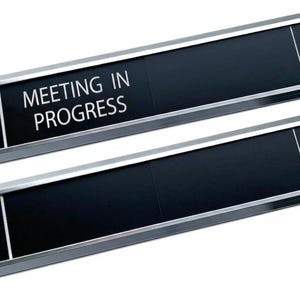 Puede incluir: Dos letreros rectangulares para puertas de oficina con fondo negro y marcos de metal plateado. El letrero superior muestra el texto blanco "MEETING IN PROGRESS" a la izquierda, mientras que el inferior está en blanco. Ambos tienen una línea.