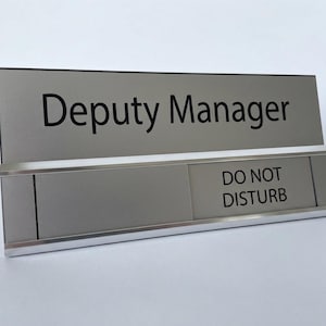 May include: A rectangular silver desk sign with a metallic finish featuring the black text Deputy Manager on the top section and a sliding panel on the bottom section that displays the black text DO NOT DISTURB in a clear, professional font.
