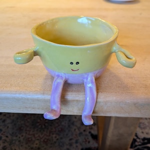 May include: A whimsical ceramic bowl with a yellow interior and exterior, featuring small loop handles and painted-on eyes and a smile. The bowl has pink legs, giving it a playful, anthropomorphic appearance.