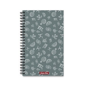 Pode incluir: Um caderno espiral cinzento com um padrão branco de símbolos abstratos. O caderno tem uma encadernação em espiral preta e um pequeno retângulo vermelho com o texto "Jason Vive" na parte inferior.