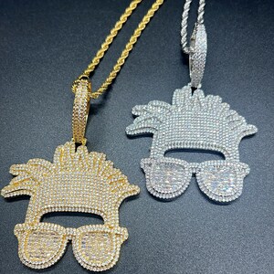 Puede incluir: Dos cadenas de oro y plata con colgantes que representan una cara de estilo de dibujos animados con gafas de sol y un peinado salvaje, todo cubierto de brillantes diamantes de imitación.