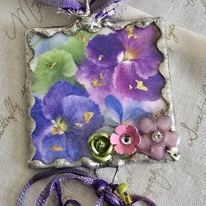 May include: A square pendant featuring a floral print of purple and green pansies with gold leaf accents, framed in silver metal. The pendant hangs from a purple ribbon and is decorated with small flower charms, beads, and dangling fabric ribbons.