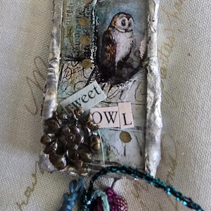 May include: A rectangular pendant with a soldered silver-toned metal frame featuring a collage of two owl illustrations, gold-toned dots, and paper cutouts with the text "sweet" and "OWL". A bronze-toned floral embellishment sits at the bottom.