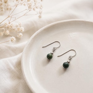 May include: A pair of dangle earrings with dark green, round beads and silver-colored hooks. The earrings are displayed on a white ceramic plate, with a soft, neutral-toned background.
