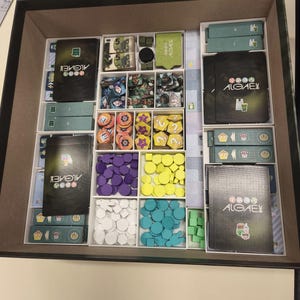 Könnte beinhalten: Geöffnete Spielbox mit sortierten Spielkomponenten. Die Box enthält Karten, Spielsteine und Spielteile in verschiedenen Farben, darunter Lila, Gelb und Blau. Der Titel des Spiels, "ALGAE", ist auf den Karten zu sehen. Die Box ist geöffnet.