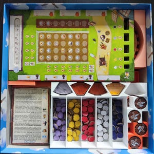 Könnte beinhalten: Ein Brettspiel in einer blauen Box mit einem farbenfrohen Spielbrett zum Thema Bauernhof, verschiedenen Spielsteinen und Anweisungskarten. Das Spielbrett zeigt ein Feldraster und einen Weg. Die Spielsteine sind in verschiedenen Farben.