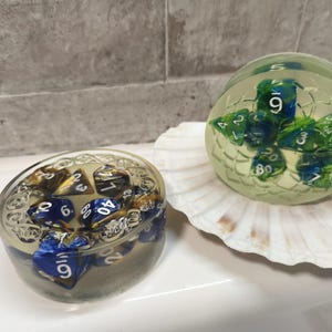 Puede incluir: Dos juegos de dados de resina. Uno es un recipiente ovalado lleno de dados azules, dorados y blancos. El otro es un recipiente esférico con dados verdes y azules, que descansa sobre una concha blanca.