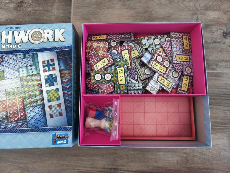 Peut inclure: Bo&icirc;te de jeu de soci&eacute;t&eacute; ouverte, intitul&eacute;e "Patchwork Nordic". La bo&icirc;te bleue clair contient des pi&egrave;ces de jeu &agrave; motifs, des boutons, un plateau de jeu et de petits &eacute;l&eacute;ments. Les pi&egrave;ces de jeu sont mesur&eacute;es en centim&egrave;tres.