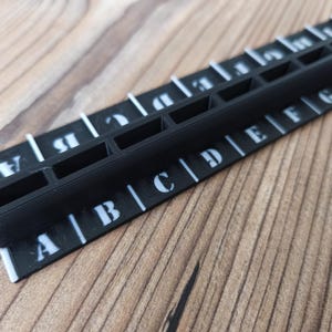 Peut inclure: Un outil noir en forme de règle avec des lettres blanches et des fentes. L'outil porte les lettres A à I. L'outil est posé sur une surface en bois.