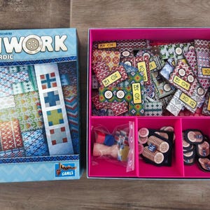 Op de afbeelding: Een bordspel genaamd "Patchwork Nordic" met een lichtblauwe doos met een patchwork-ontwerp. De doos is open en onthult een roze bak gevuld met spelonderdelen, waaronder stoffen tegels, knopen en andere componenten.