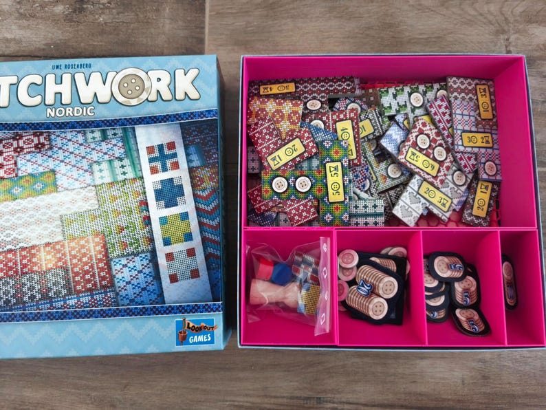 Peut inclure: Jeu de soci&eacute;t&eacute; "Patchwork Nordic" avec une bo&icirc;te bleue clair orn&eacute;e d'un motif patchwork. La bo&icirc;te ouverte r&eacute;v&egrave;le un plateau rose rempli de pi&egrave;ces de jeu, notamment des tuiles en tissu, des boutons et d'autres composants.