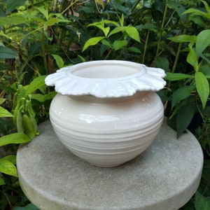 May include: White ceramic planter with a ruffled rim. The planter has a smooth, white glaze and a slightly rounded shape.