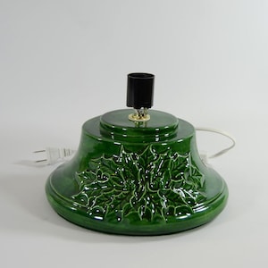 May include: Green ceramic Christmas tree stand with a holly leaf design. The stand has a black light socket and a white cord.