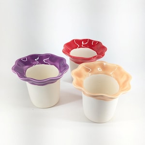 Puede incluir: Tres macetas de cerámica con bordes con volantes en morado, rojo y melocotón. Las macetas tienen una base blanca y están dispuestas sobre una superficie blanca. Ideales para plantas pequeñas o decoración del hogar.