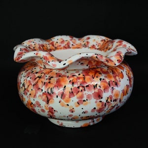 May include: A ceramic bowl with a ruffled rim, decorated with a white base and a pattern of orange, red, and black floral-like spots. The bowl has a rounded body and a pedestal base, set against a black background.