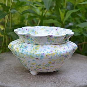May include: A white ceramic planter with a speckled glaze in shades of yellow, green, and purple. The planter has a wide, scalloped rim and three small feet.