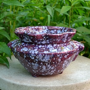 May include: A purple and white speckled ceramic planter with a scalloped rim and three legs. The planter has a saucer for drainage.