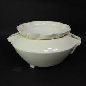 May include: A cream-colored ceramic serving dish with a matching lid. The dish has a rounded base with three small feet and two handles. The lid is a shallow, scalloped-edged plate. The set is likely for serving food.
