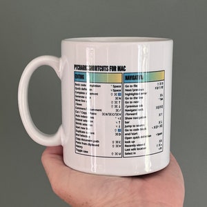 May include: White ceramic mug with black text listing PyCharm shortcuts for Mac. The mug has a blue, yellow, and green color scheme. The text includes keyboard shortcuts for editing and navigating code.