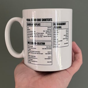 May include: White ceramic mug with black text listing Visual Studio Code shortcuts for search and replace, multi-cursor selection, and file management/terminal.