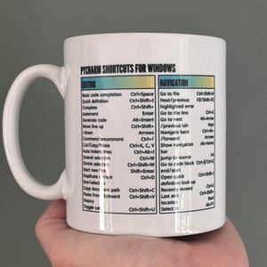 May include: White ceramic mug featuring "PYCHARM SHORTCUTS FOR WINDOWS" text. The mug displays keyboard shortcuts for editing and navigation, with a handle on the left. The text is in black, yellow, and green.