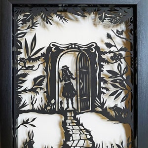 May include: Black and white paper cut art depicting a girl in a dress and hat standing in front of a doorway with a path leading away from it. The artwork is framed in black.