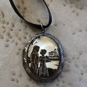 May include: A silver oval locket with a black ribbon. The locket has a glass front with a silhouette image of a couple standing in front of a building.