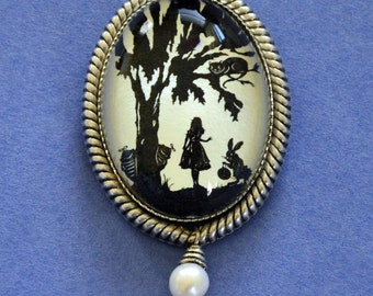 May include: A silver oval brooch with a white cameo featuring a silhouette of Alice in Wonderland standing in a forest with a cat and a rabbit. The brooch has a pearl drop pendant.