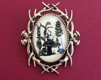 May include: A silver-toned brooch with a glass oval cameo featuring a black silhouette of Alice in Wonderland sitting on a mushroom with the Mad Hatter. The cameo is surrounded by a decorative antler frame.