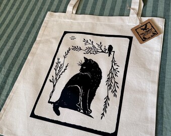 May include: A white tote bag with black handles and a black silhouette print of a cat sitting in a field of grass with a bird perched on a branch. The text "The Crafty Raven" is printed on a brown tag attached to the bag.
