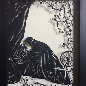 May include: Black and white paper cut-out art depicting a couple embracing in a forest setting. The woman is wearing a dress and the man is wearing a cloak. A spinning wheel is visible in the background.
