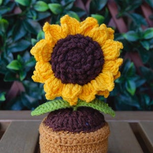 May include: A crocheted sunflower in a pot. The sunflower features bright yellow petals and a dark brown centre. The pot is light brown, and the leaves are green. The item is placed on a wooden surface.