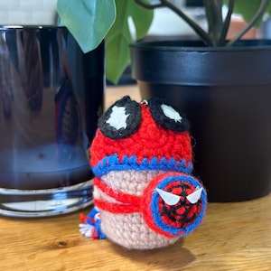 May include: A handmade crochet Spider-Man doll. The doll features a red and blue color scheme, with black and white accents. The doll has a round shape and a small, circular shield with a spider web design.