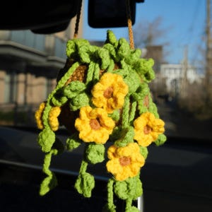 Pode incluir: Uma cesta suspensa de crochê feita à mão com flores amarelas e folhas verdes. A cesta é marrom e suspensa por um cordão marrom. As flores têm centros marrons. O fundo é uma visão desfocada de um dia ensolarado.