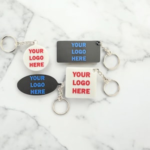May include: Three different keychains with the text "YOUR LOGO HERE" in red and blue lettering. The keychains are made of black, white, and gray plastic.