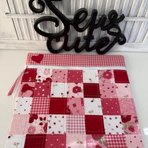 May include: A patchwork zippered pouch in shades of red, pink, and white. The design features squares with hearts, flowers, and gingham patterns. The top edge has a red and white gingham trim. A black cursive sign that says "Sew Cute" is above the pouch.