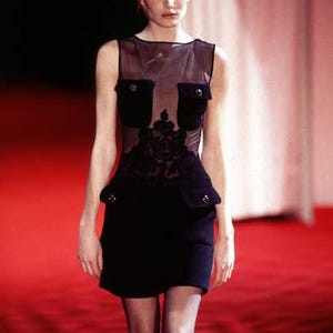 May include: A woman wearing a black sheer dress with a floral design and a black skirt. The dress has two pockets on the front. She is standing on a red carpet.