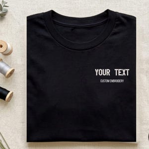 May include: A black crew neck t-shirt featuring white embroidered text on the right chest area. The text reads "YOUR TEXT" in a larger font size, with the words "CUSTOM EMBROIDERY" printed in a smaller font size directly underneath the main text.