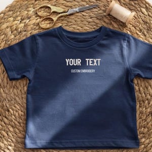 May include: A navy blue short-sleeved t-shirt featuring white embroidered text that reads YOUR TEXT and CUSTOM EMBROIDERY centred on the chest. The shirt is displayed with gold-toned metal scissors and a spool of thread nearby.