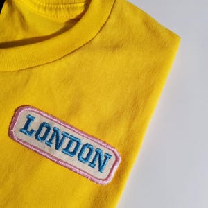 May include: A bright yellow t-shirt featuring a rectangular embroidered patch on the chest. The patch has a cream-coloured background with a light pink border and the word LONDON stitched in bold, blue, capital letters across the centre.
