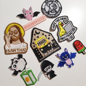 Könnte beinhalten: Eine Sammlung von gestickten Aufnähern, darunter eine rosa Fledermaus, ein runder Hundeaufnäher, eine Ente am Computer mit Text, eine Jesus-Figur mit dem Text I SAW THAT, ein Bus mit dem Text MASR, ein Panda, ein Sensenmann, eine blaue Fledermaus und ein Eis am Stiel.